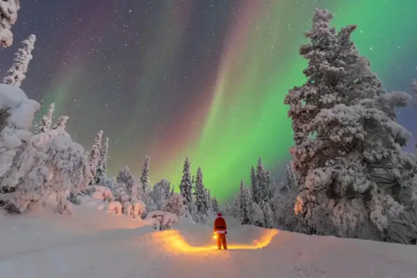 Un uomo con lanterna, vestito da Babbo Natale, ammira l'aurora boreale in Lapponia. Un paesaggio innevato e magico, perfetto per chi cerca dove andare in Lapponia.